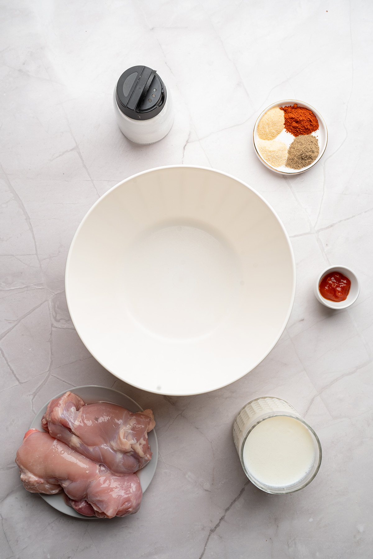 Ingredients for buttermilk fried chicken sandwich arranged on a counter, including raw chicken thighs, buttermilk, spices, salt, and hot sauce.