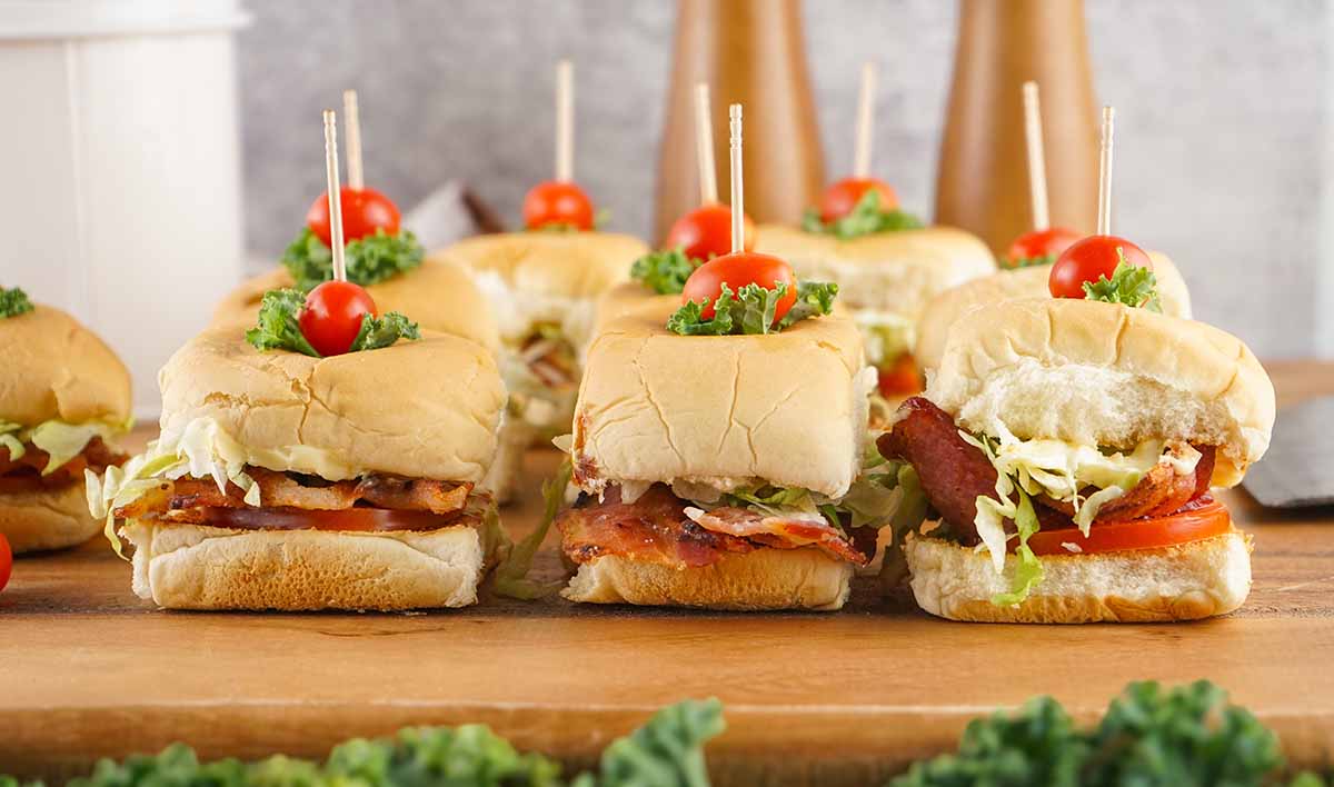 Front view of a row of BLT sliders, showing all the layers and garnishes on a wooden board