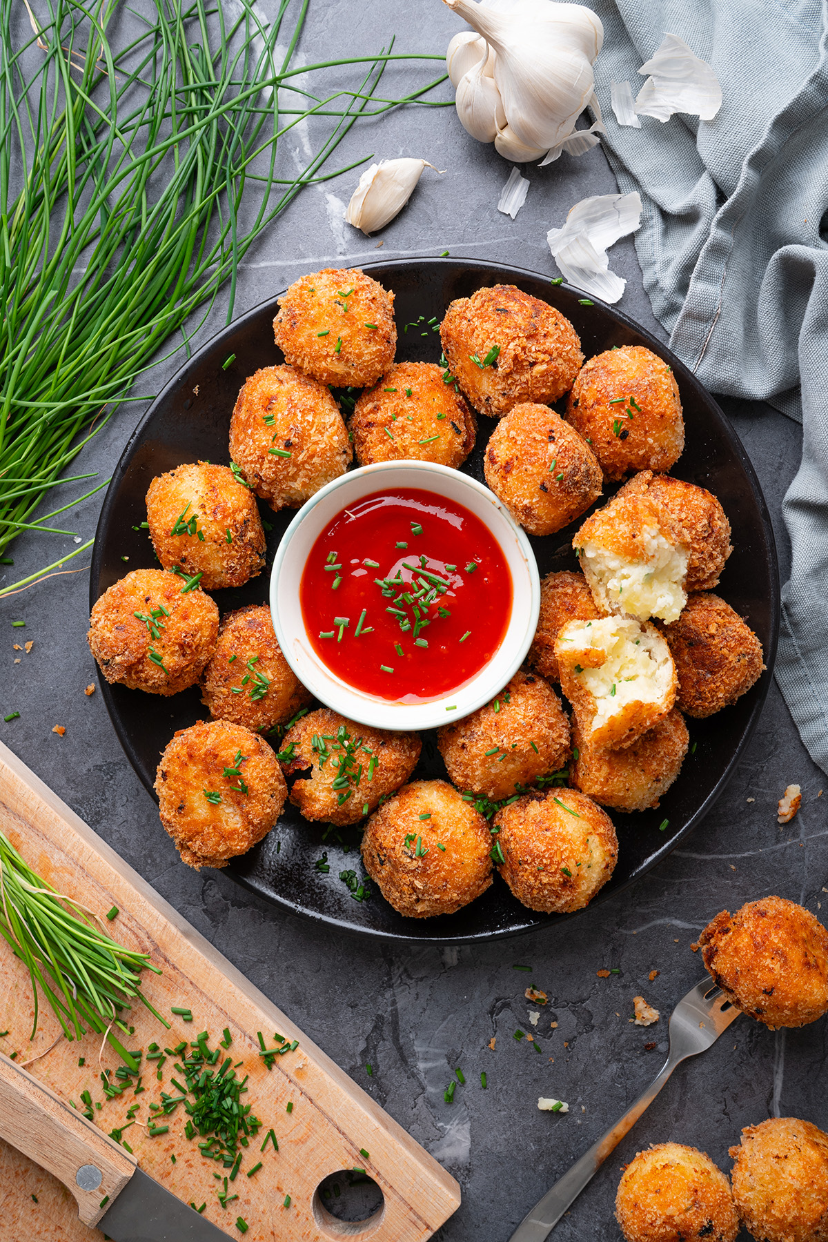 These fried mashed potato balls are crispy, golden, and loaded with cheesy flavor. The perfect way to turn leftover mashed potatoes into an irresistible side or snack. Overhead view of golden, crispy fried mashed potato balls arranged on a black plate, garnished with chopped chives and served with a bowl of red dipping sauce in the center.