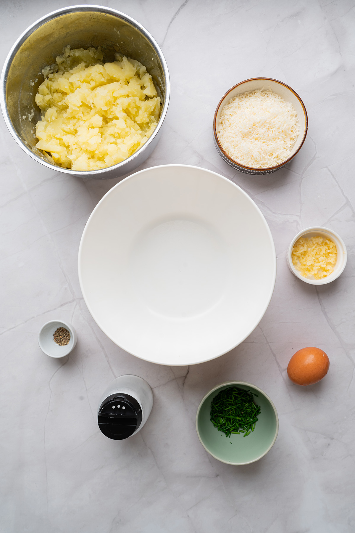These fried mashed potato balls are crispy, golden, and loaded with cheesy flavor. The perfect way to turn leftover mashed potatoes into an irresistible side or snack. Top-down image of individual ingredients for fried mashed potato balls including mashed potatoes, grated Parmesan, chopped chives, garlic, an egg, salt, pepper, and an empty mixing bowl.