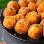 Close-up shot of golden fried mashed potato balls piled high on a black plate, with crispy panko coating and fresh herbs sprinkled on top.