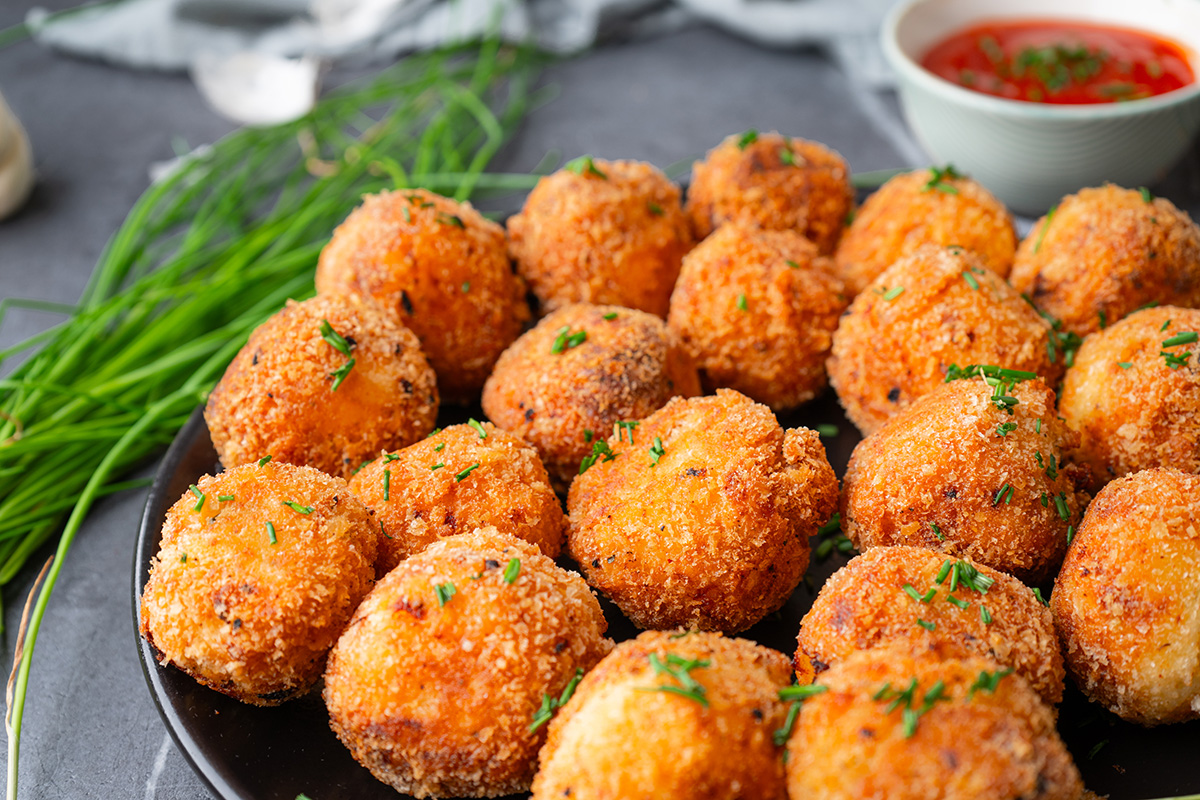 These fried mashed potato balls are crispy, golden, and loaded with cheesy flavor. The perfect way to turn leftover mashed potatoes into an irresistible side or snack. Angled view of a full plate of fried mashed potato balls, showing their crispy texture and garnished with chopped chives. A bowl of red dipping sauce sits in the background.