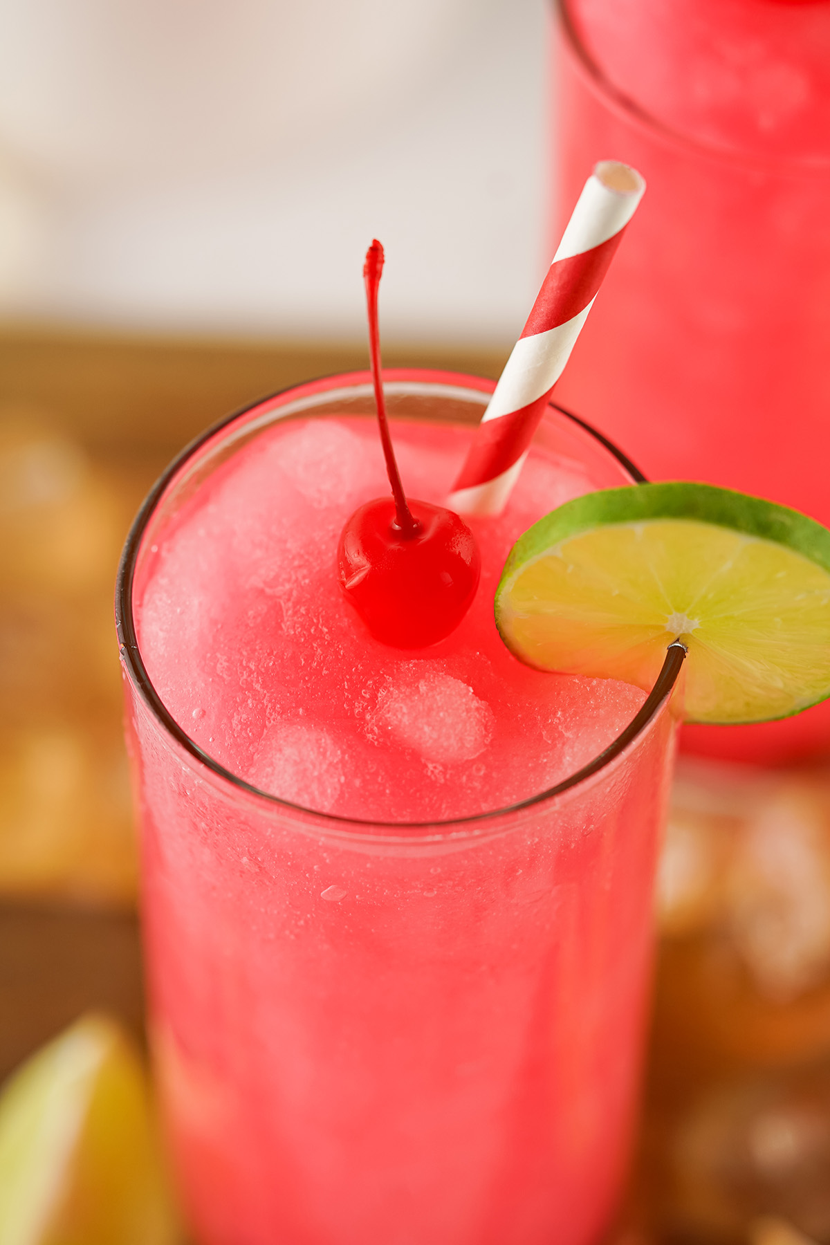 Bright pink slushy Dirty Shirley cocktail topped with a maraschino cherry and lime slice, served with a red and white striped straw in a tall glass.