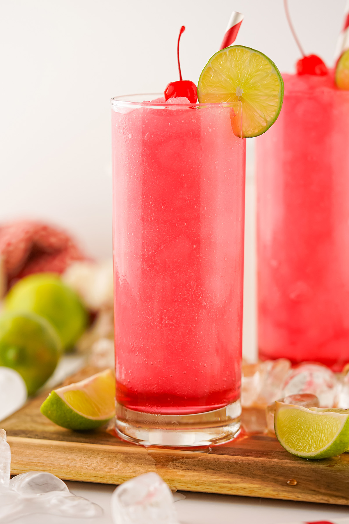 Two tall Frozen Dirty Shirley cocktails on a wooden tray, each garnished with a lime slice, cherry, and paper straw, with scattered ice cubes and fresh limes in the background.
