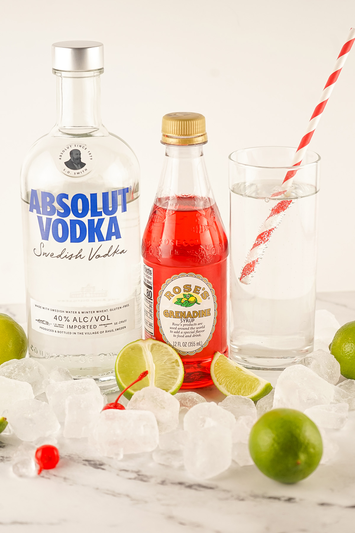 Bottles of Absolut vodka and Rose’s grenadine next to a glass of lemon-lime soda, surrounded by ice cubes, lime wedges, and maraschino cherries.