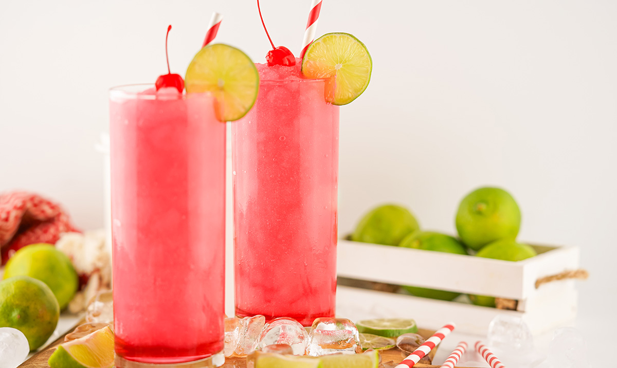 Tall glass filled with a vibrant pink Frozen Dirty Shirley, garnished with a cherry and lime wheel, surrounded by ice and lime wedges on a wooden board.