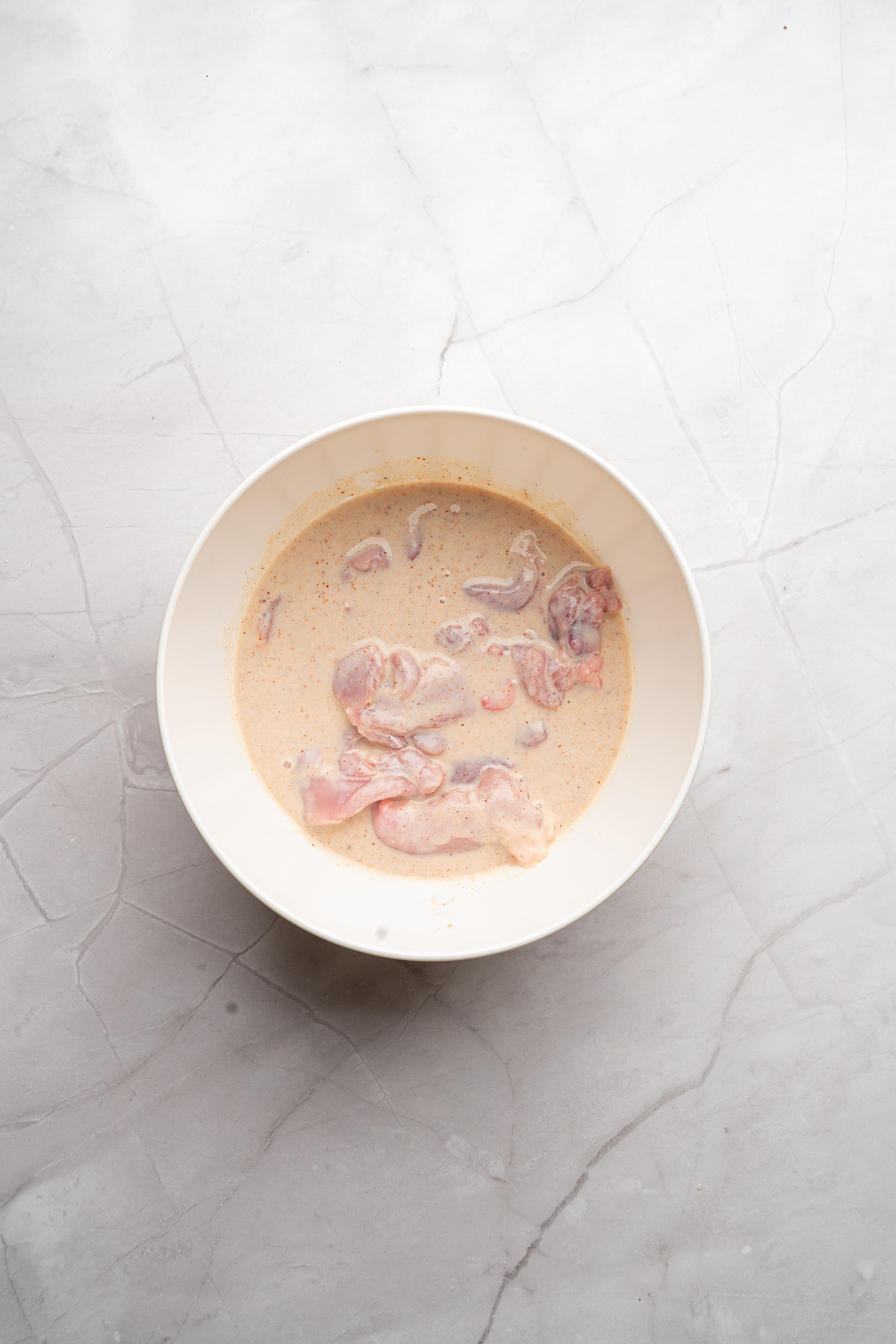 Raw chicken pieces soaking in a seasoned buttermilk marinade inside a large white bowl.