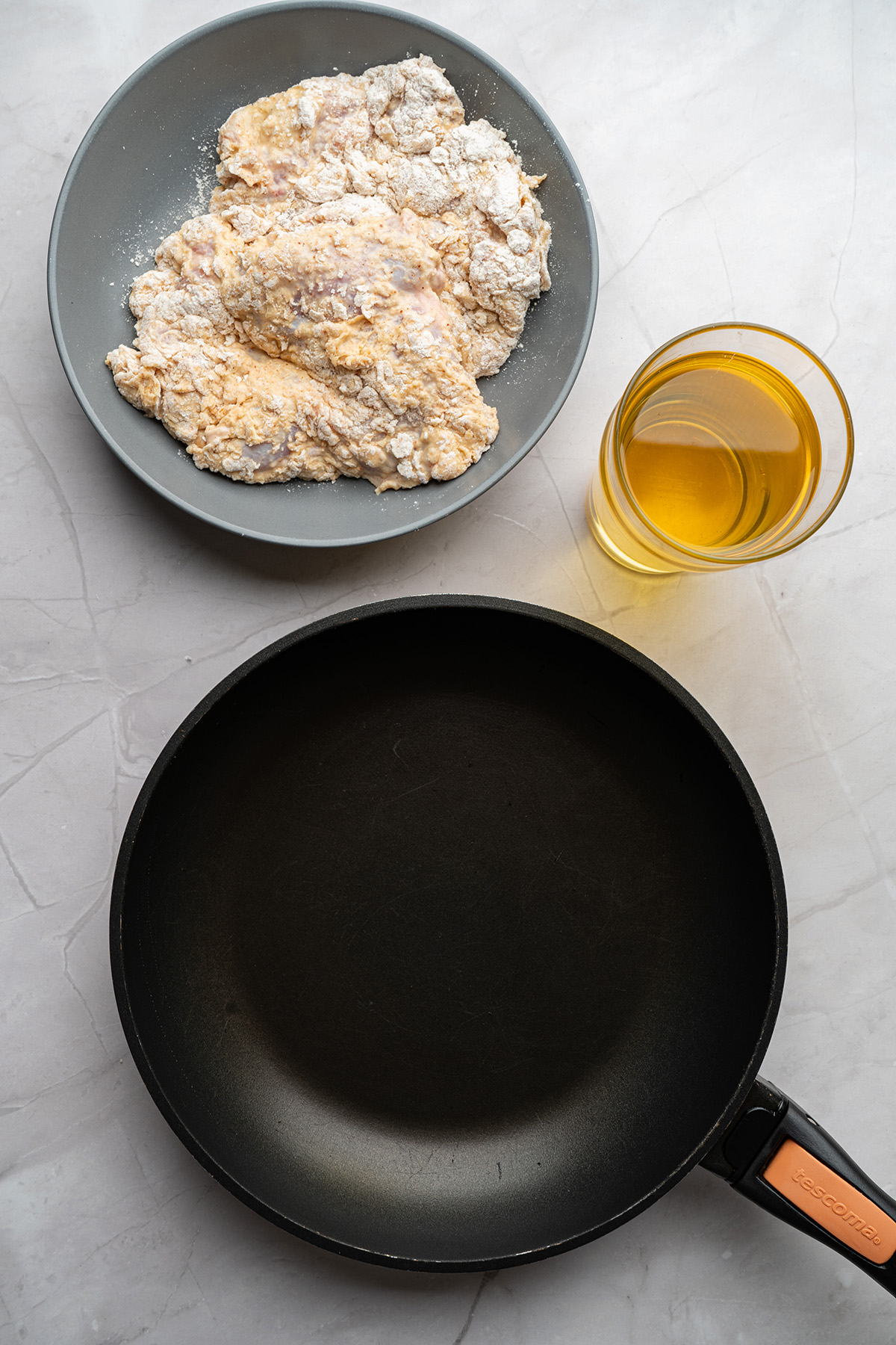 Breaded chicken pieces resting before frying with a glass of oil and skillet nearby.