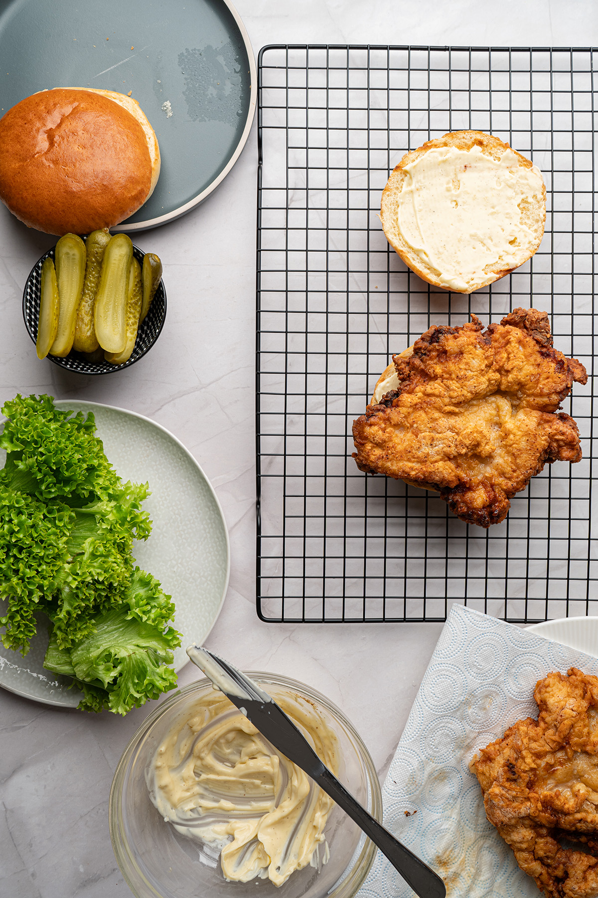 Fried chicken sandwich assembly on a wire rack with crispy chicken, toasted buns, pickles, lettuce, and mayonnaise.