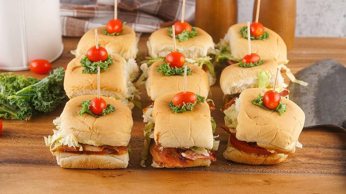 Assembled BLT sliders on a wooden board, each secured with a toothpick, cherry tomato, and small kale leaf garnish.