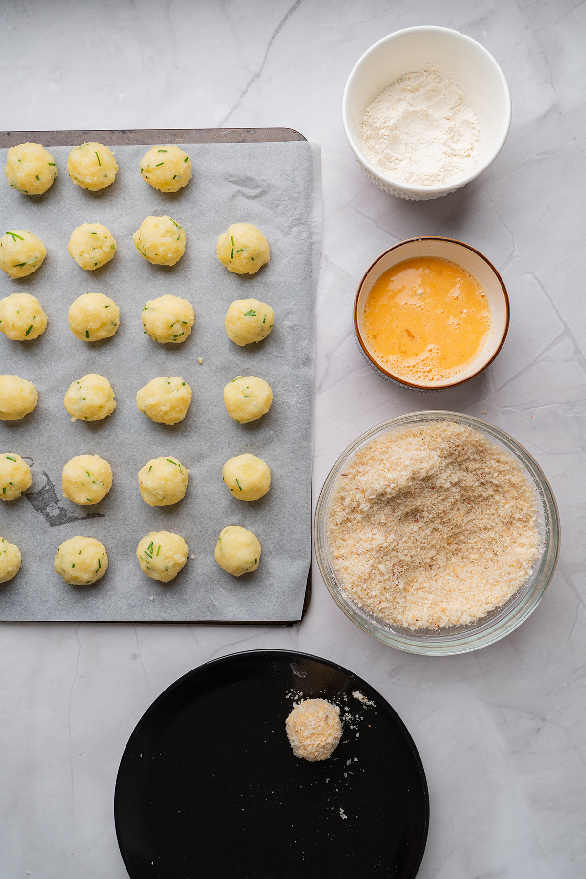 These fried mashed potato balls are crispy, golden, and loaded with cheesy flavor. The perfect way to turn leftover mashed potatoes into an irresistible side or snack. Step-by-step prep image showing shaped mashed potato balls on a parchment-lined tray next to bowls of flour, beaten eggs, and seasoned breadcrumbs.