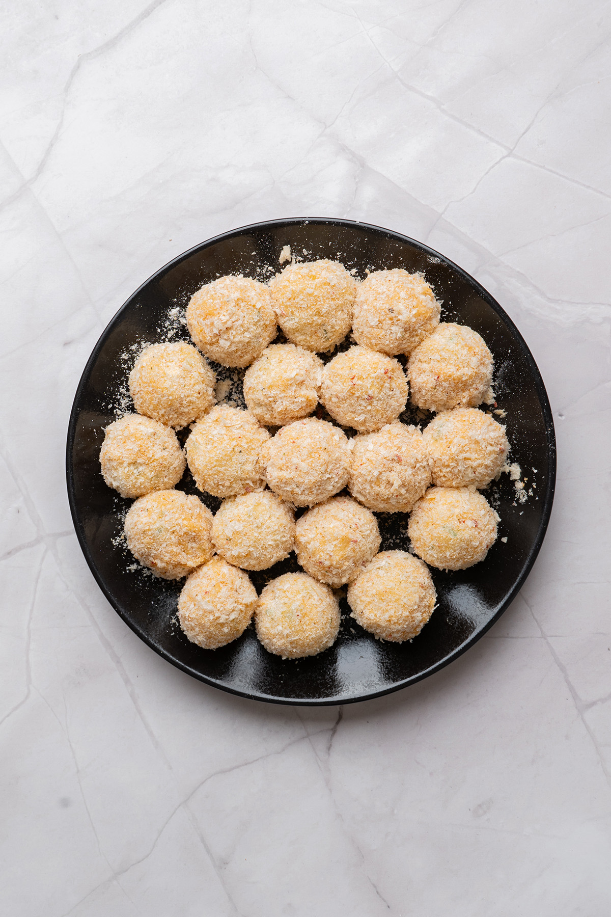 These fried mashed potato balls are crispy, golden, and loaded with cheesy flavor. The perfect way to turn leftover mashed potatoes into an irresistible side or snack. Overhead image showing mashed potato balls coated in breadcrumbs and neatly arranged on a black plate, ready for frying.