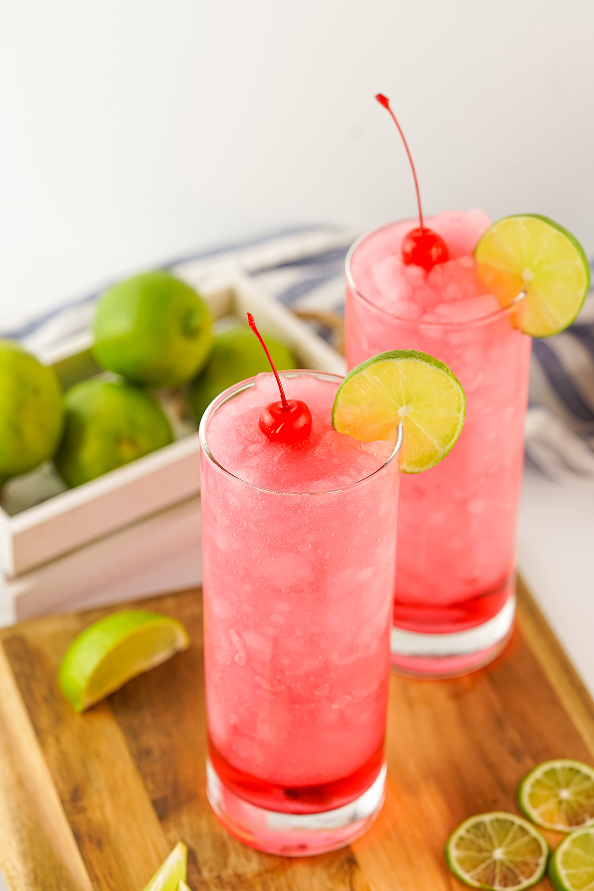 Two Frozen Dirty Shirley cocktails on a wooden serving board, garnished with cherries and lime slices, with fresh limes and a striped napkin in the background.