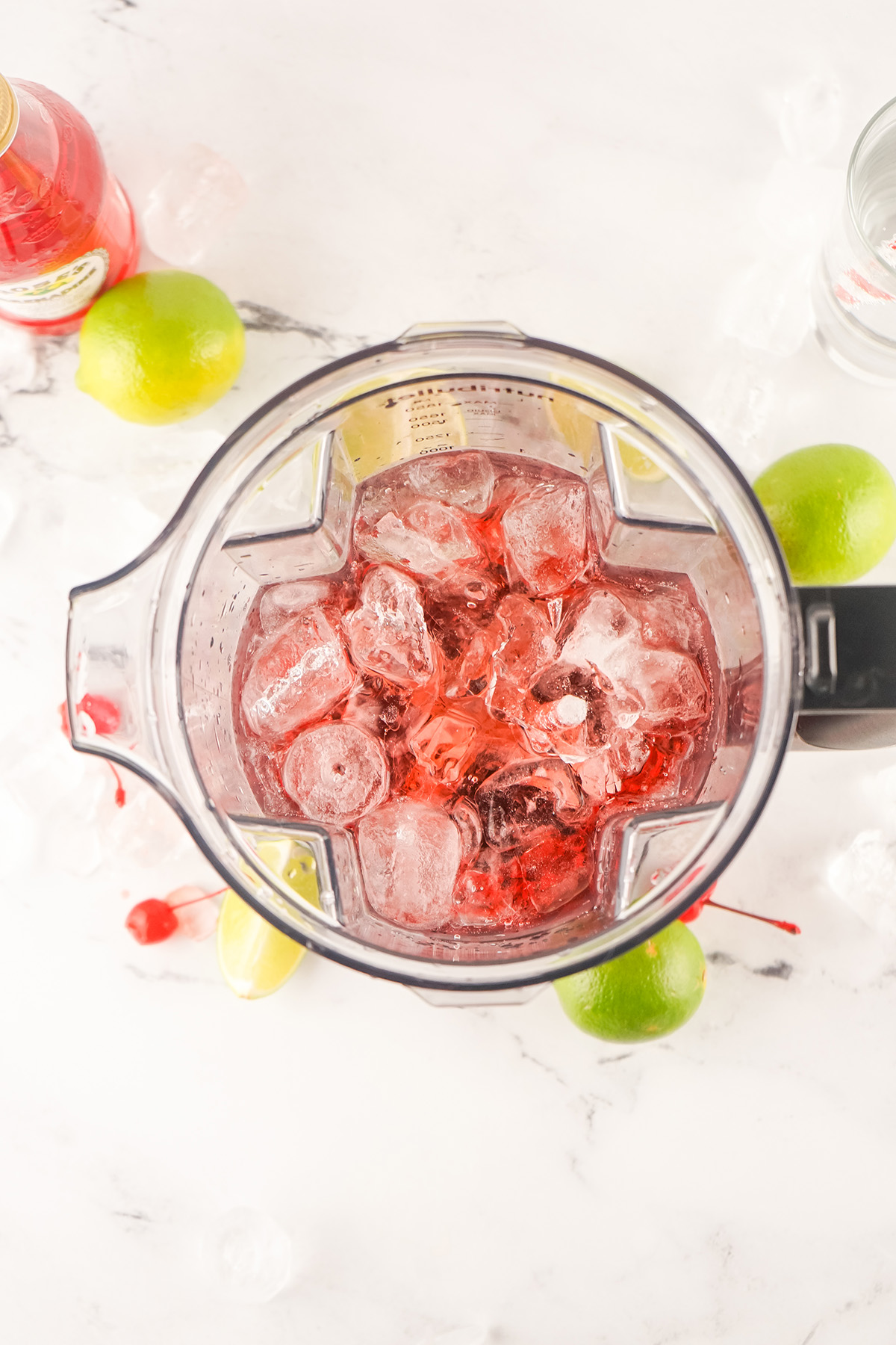 Top-down view of a blender filled with ice, grenadine, and soda, ready to blend into a Frozen Dirty Shirley, surrounded by limes and cherries on a marble surface.