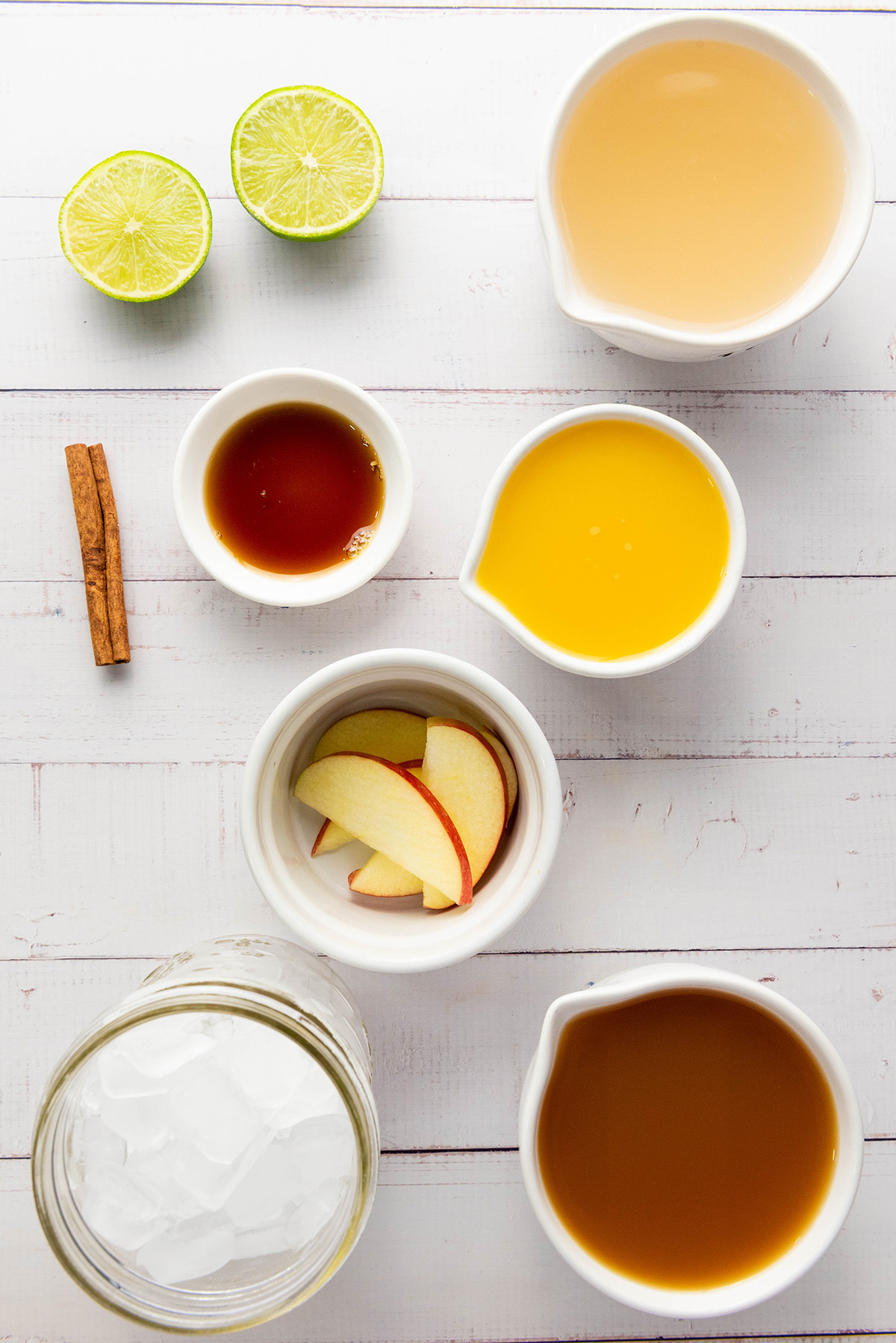 Overhead view of apple cider mocktail ingredients on a white background, including apple cider, orange juice, lime, cinnamon stick, honey, apple slices, and a glass jar of ice.