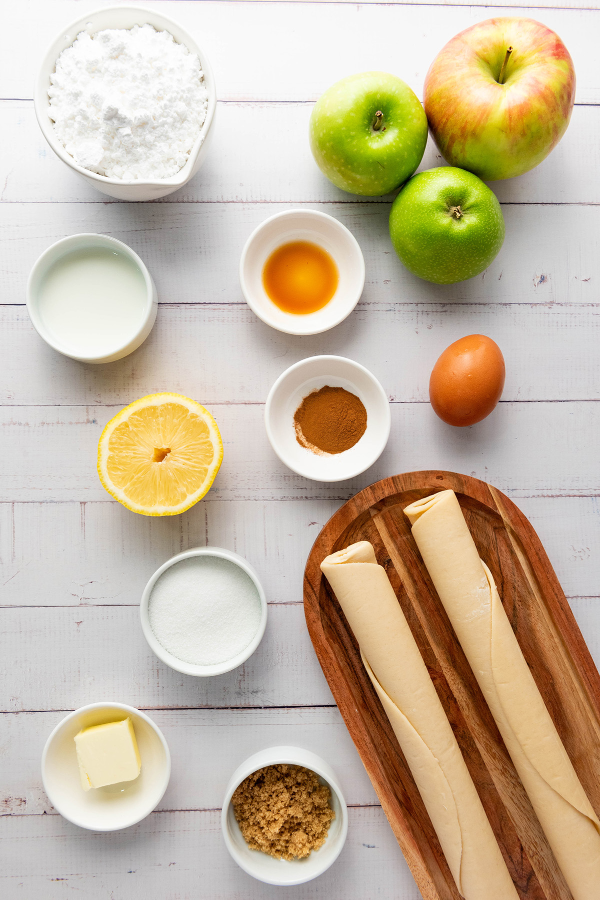 Cozy up with these easy apple hand pies made with flaky store-bought crust and a sweet cinnamon filling. All the comfort of homemade apple pie in an easy grab-and-go treat. Ingredients for apple hand pies including apples, lemon, sugar, butter, cinnamon, egg, pie dough, and powdered sugar arranged on a white table.