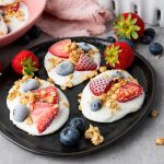 Black plate of frozen Greek yogurt bark with strawberries, blueberries, and granola, surrounded by fresh strawberries, blueberries, and a pink napkin.