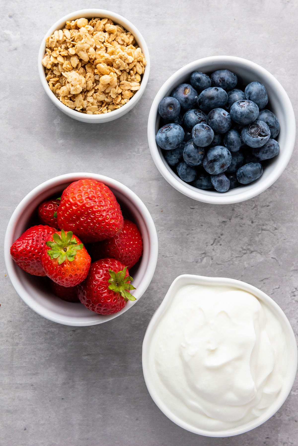 Cool, creamy, and topped with fresh fruit and crunchy granola, this frozen Greek yogurt bark is an easy, healthy treat you can make in minutes. Overhead view of small bowls holding granola, blueberries, strawberries, and Greek yogurt on a gray surface.
