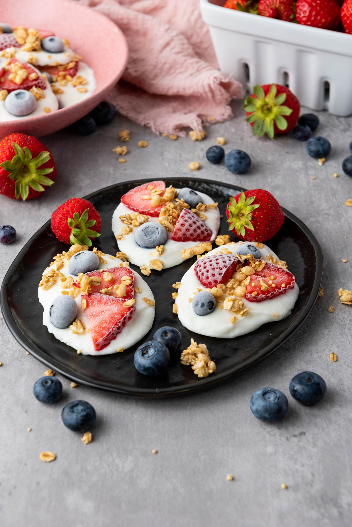 Black plate with frozen Greek yogurt bark topped with strawberries, blueberries, and granola, with fresh berries scattered around.