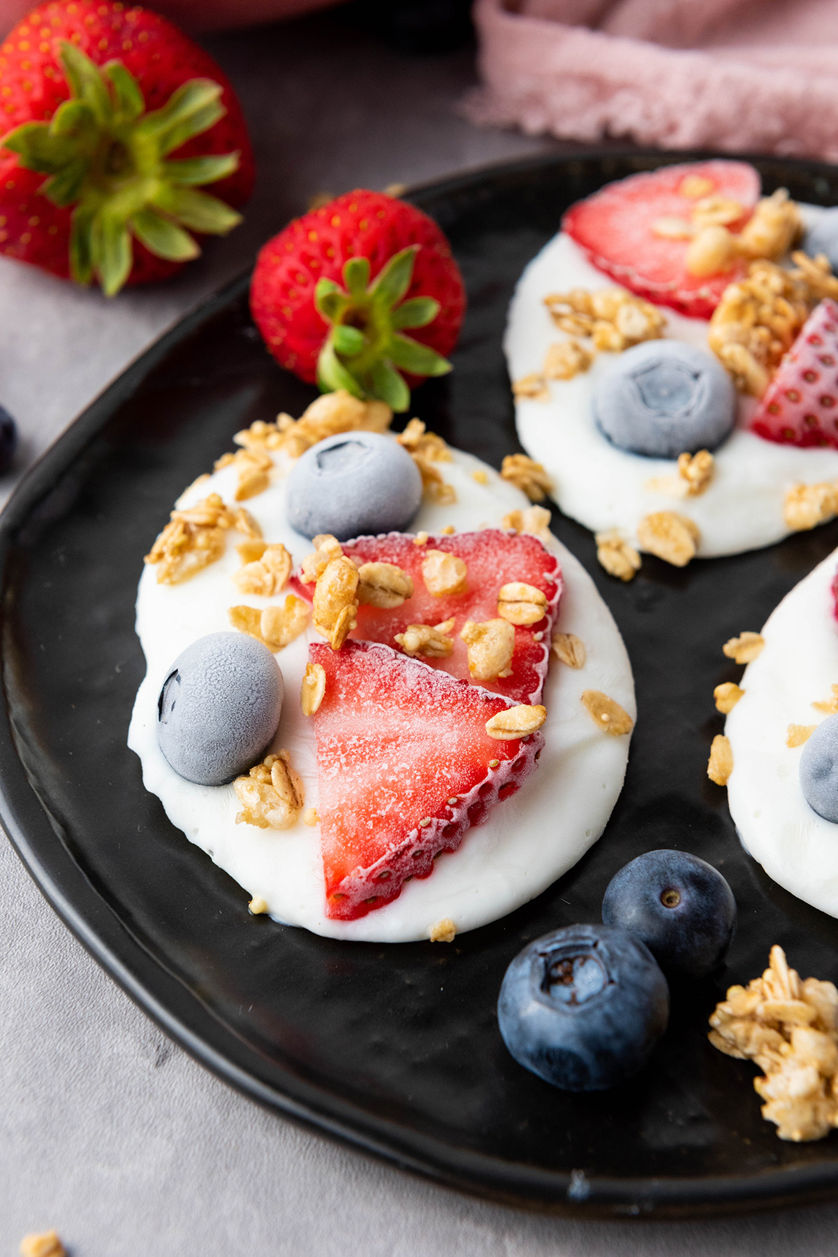 Cool, creamy, and topped with fresh fruit and crunchy granola, this frozen Greek yogurt bark is an easy, healthy treat you can make in minutes. Close-up of a frozen Greek yogurt bark topped with strawberry slices, blueberries, and granola on a black plate with fresh berries in the background.