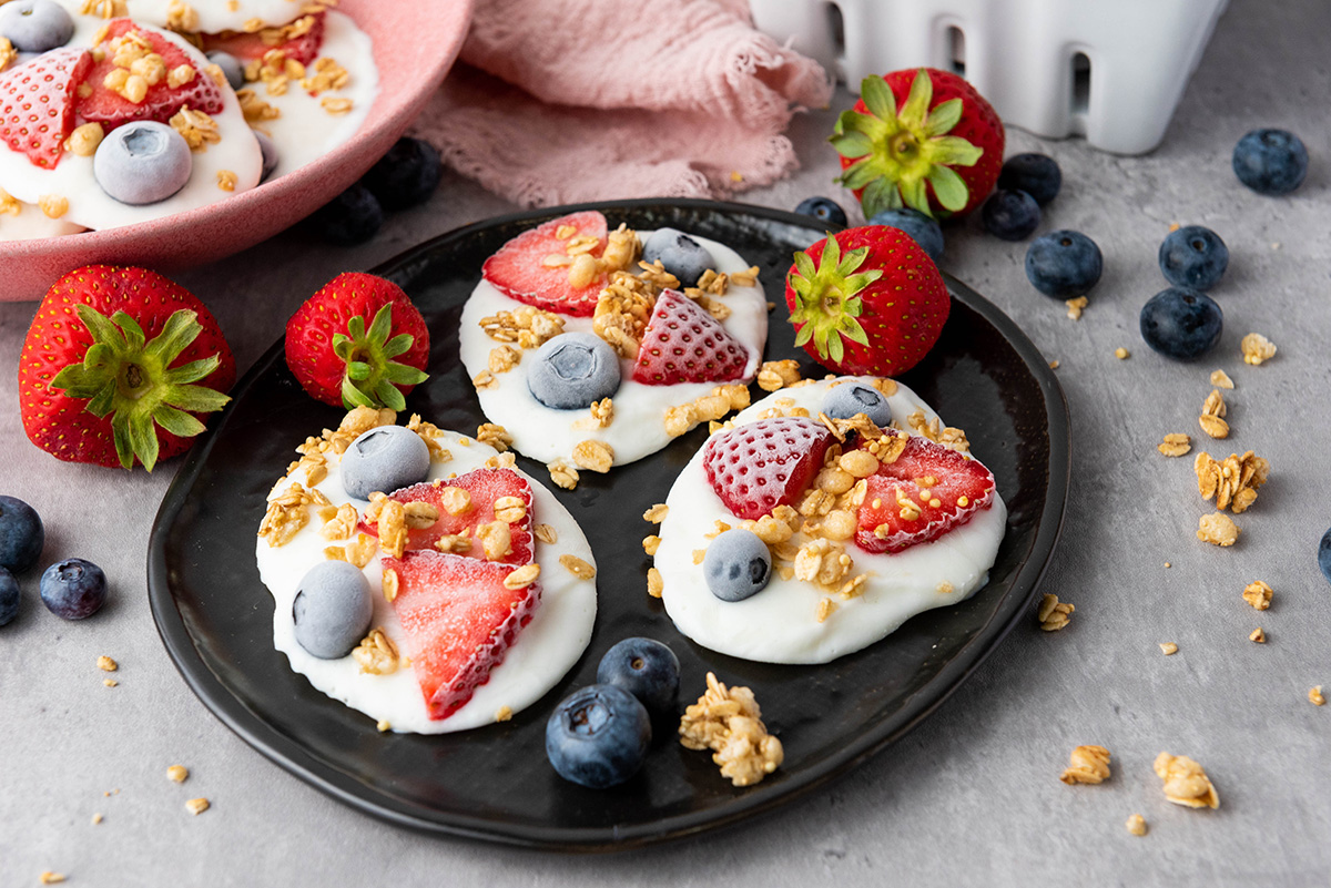 Cool, creamy, and topped with fresh fruit and crunchy granola, this frozen Greek yogurt bark is an easy, healthy treat you can make in minutes. Black plate of frozen Greek yogurt bark with strawberries, blueberries, and granola, surrounded by fresh strawberries, blueberries, and a pink napkin.