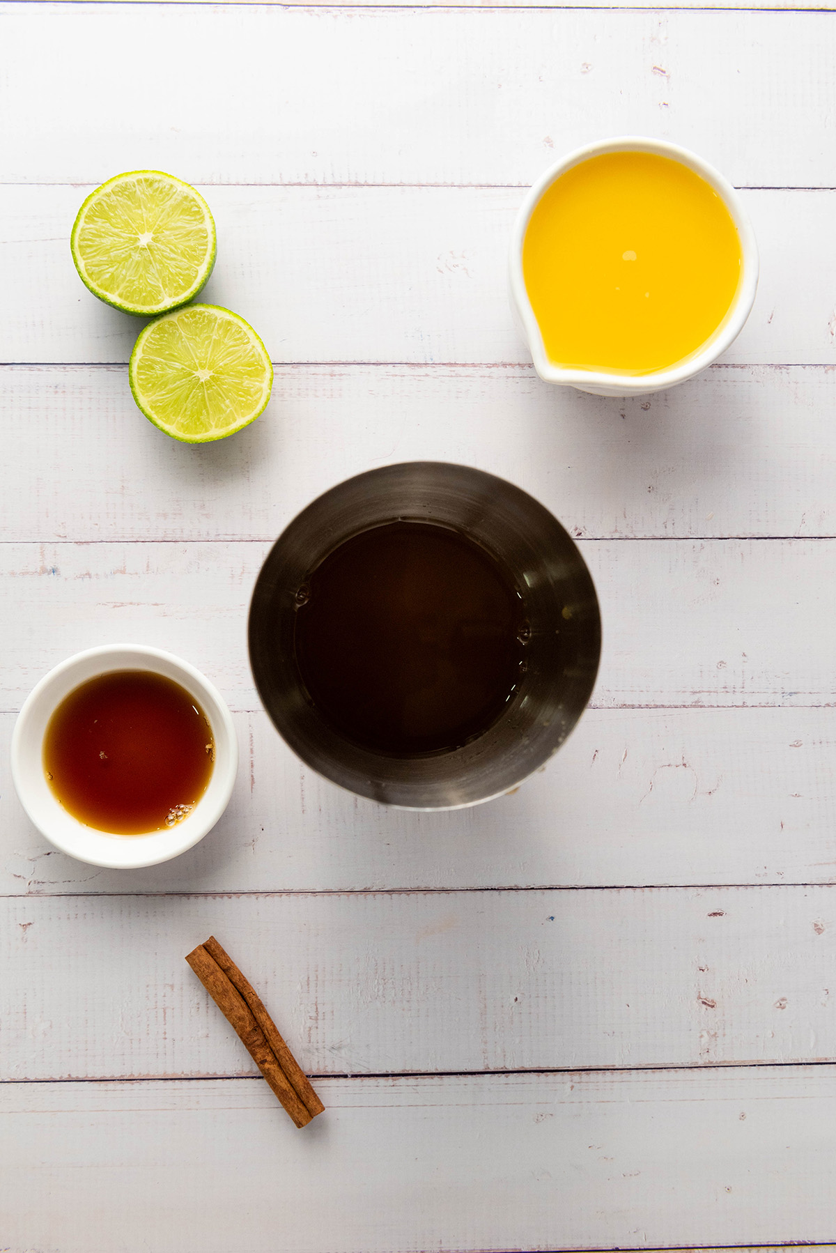 Fresh lime halves, orange juice, honey, cinnamon stick, and apple cider in a mixing tin on a white surface, ready to combine for the mocktail.
