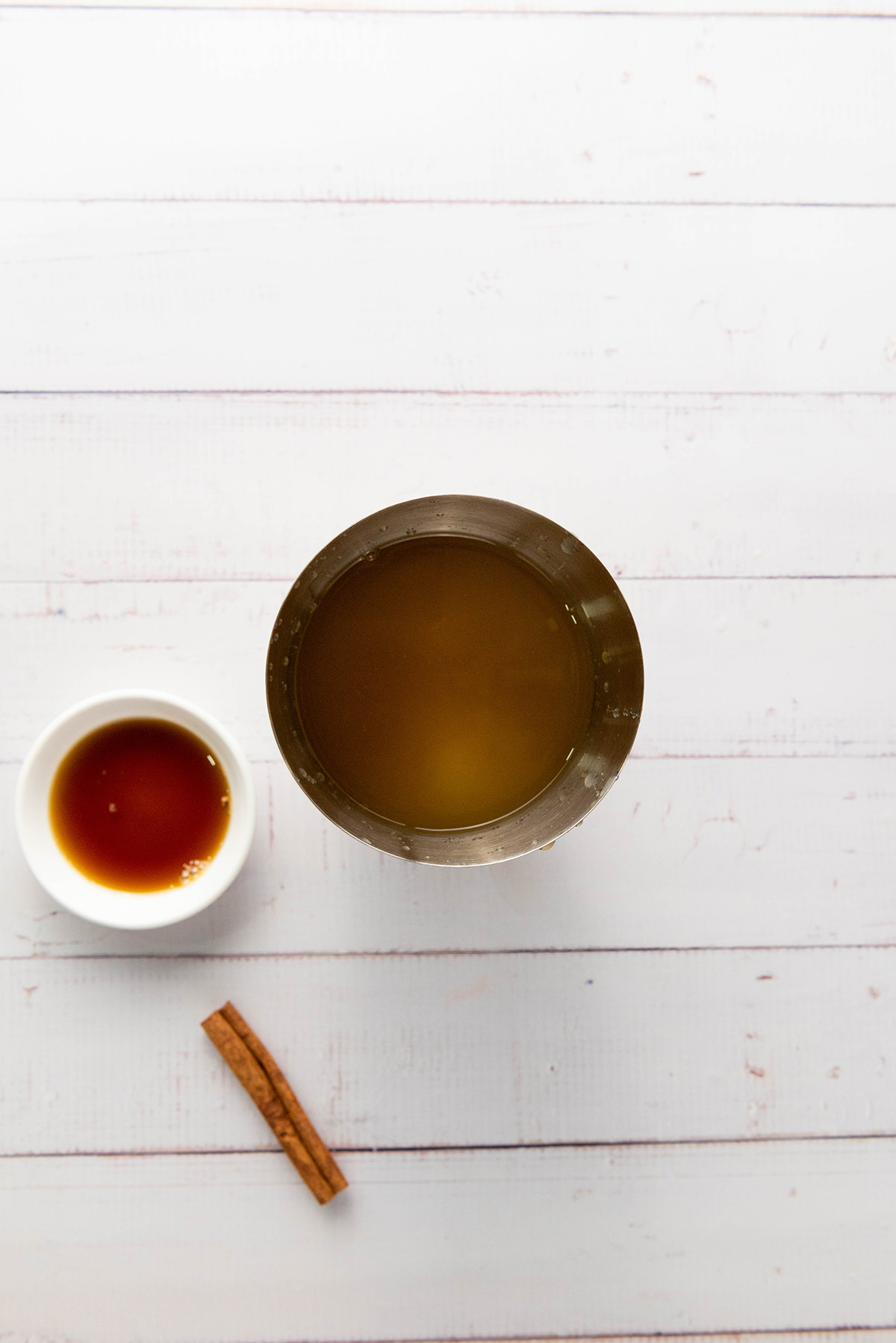 Mixing tin filled with apple cider mixture alongside honey and a cinnamon stick on a white background.