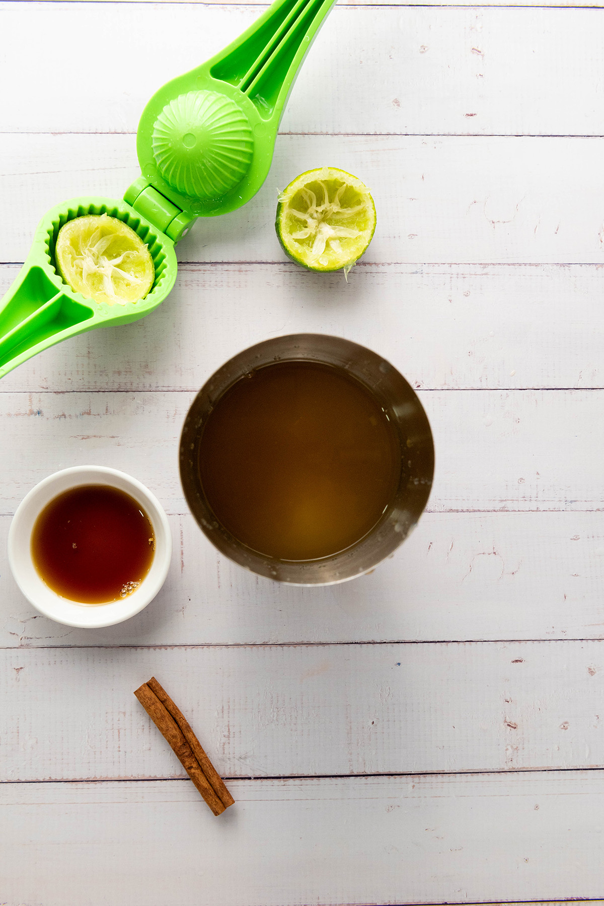 Juiced lime in a green hand juicer with apple cider mixture, honey, and cinnamon stick arranged on a white surface.