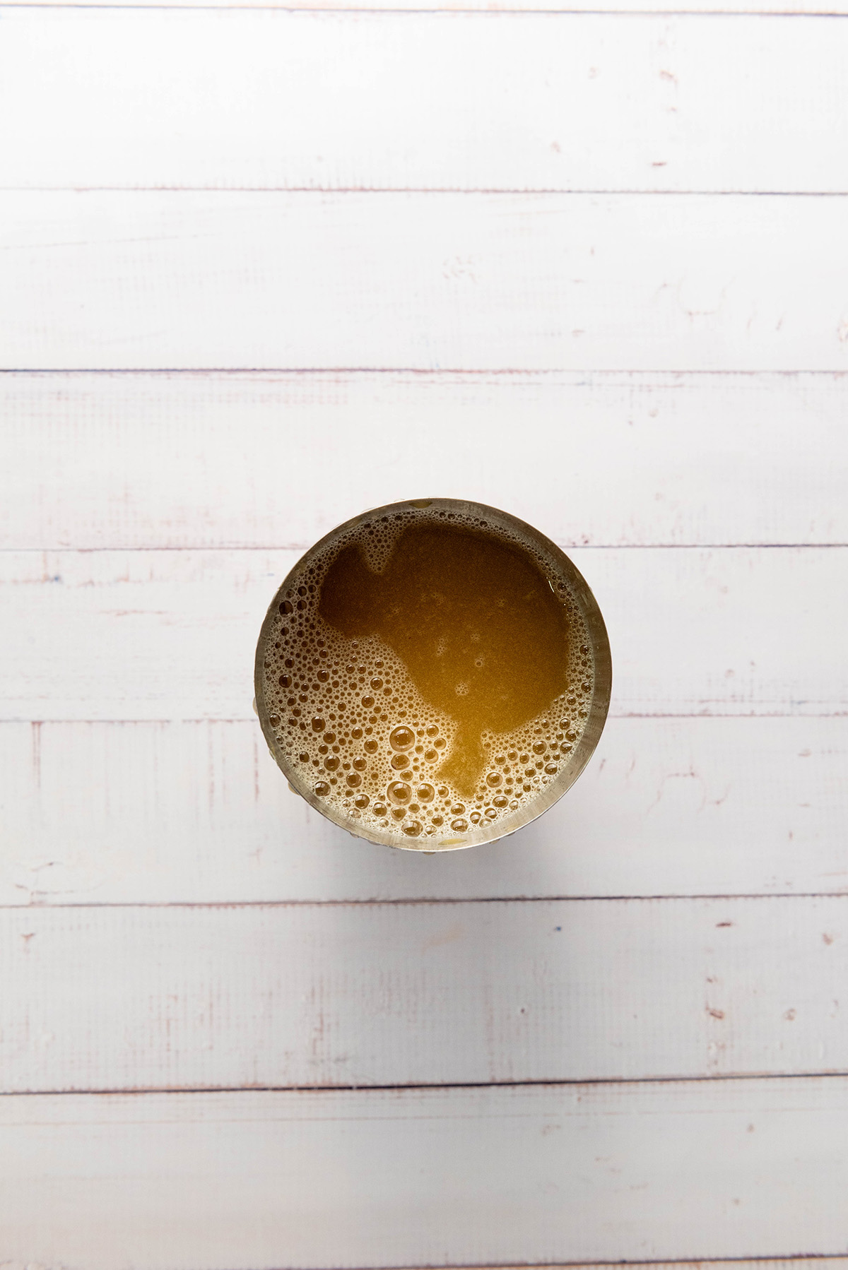 Overhead view of a mixing tin filled with the apple cider mocktail base after shaking, showing bubbles and froth on top before serving.