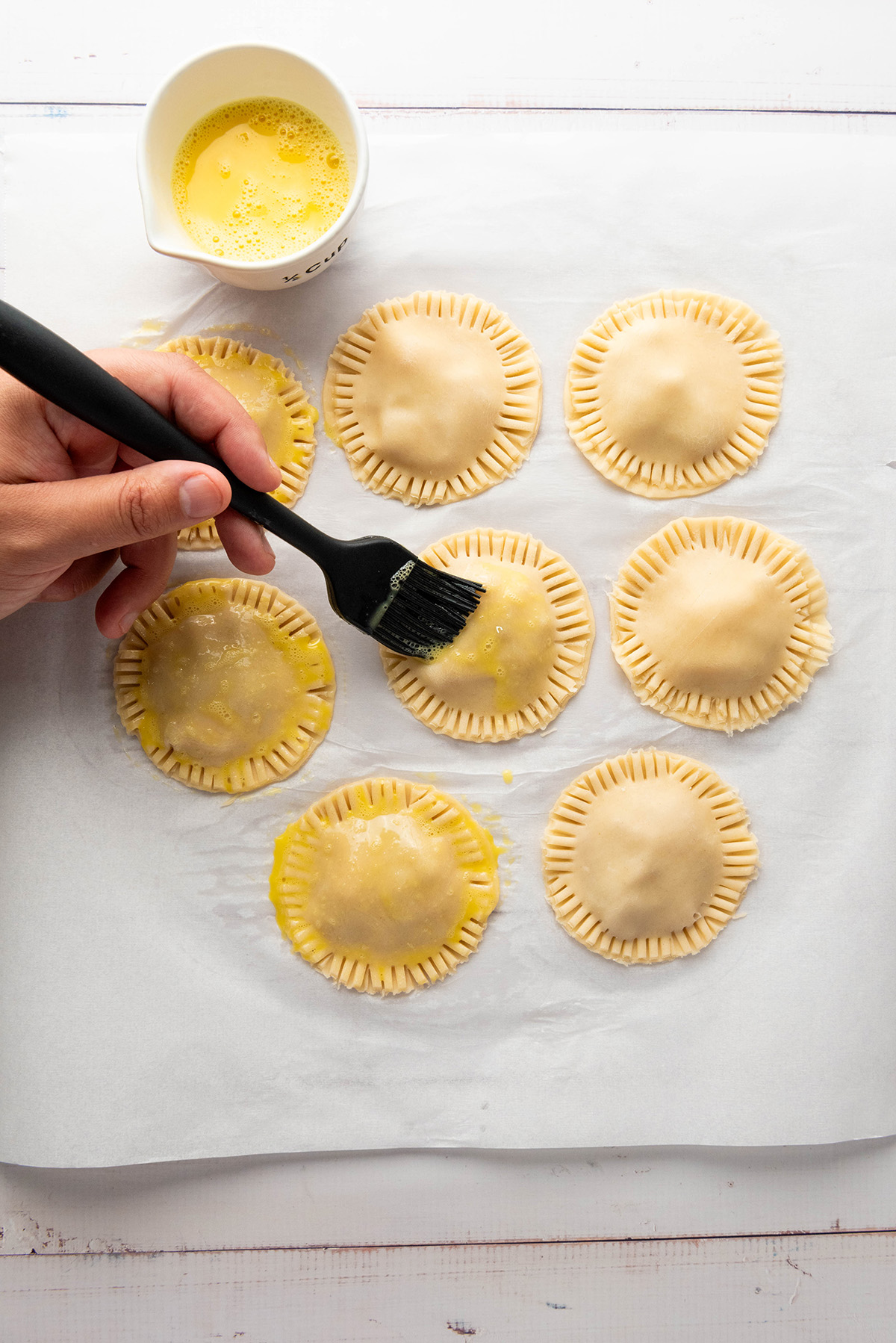 Cozy up with these easy apple hand pies made with flaky store-bought crust and a sweet cinnamon filling. All the comfort of homemade apple pie in an easy grab-and-go treat. Hand brushing egg wash over unbaked apple hand pies with crimped edges before baking.