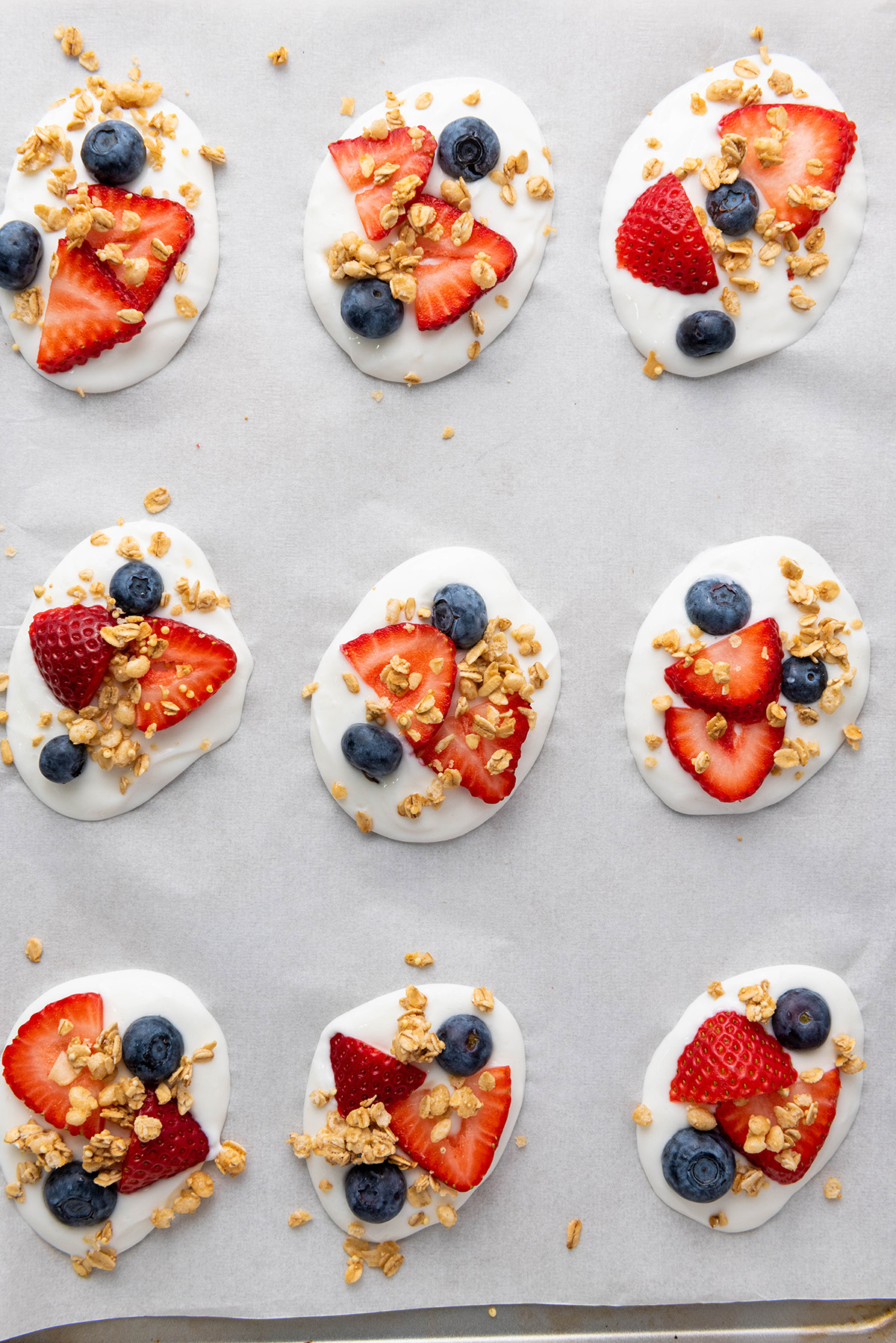 Cool, creamy, and topped with fresh fruit and crunchy granola, this frozen Greek yogurt bark is an easy, healthy treat you can make in minutes. Close-up of frozen Greek yogurt bites topped with sliced strawberries, blueberries, and granola on a parchment-lined baking sheet.