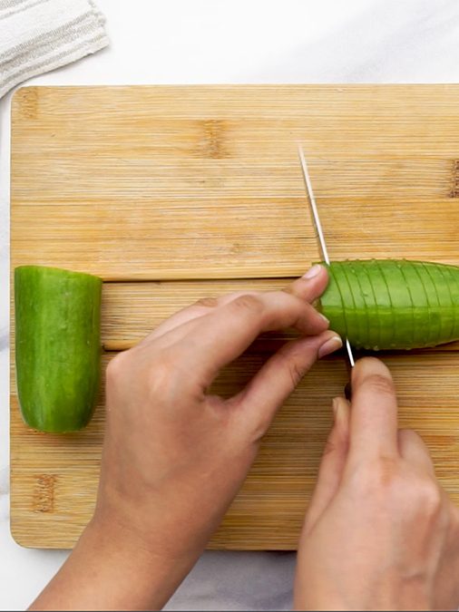 Hands slicing a cucumber placed between two chopsticks on a wooden cutting board, with garlic cloves and sesame seeds nearby.