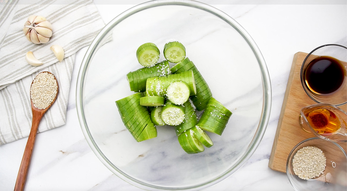 Sliced cucumbers sprinkled with salt in a glass mixing bowl, surrounded by sesame seeds, soy sauce, garlic, and chili oil.