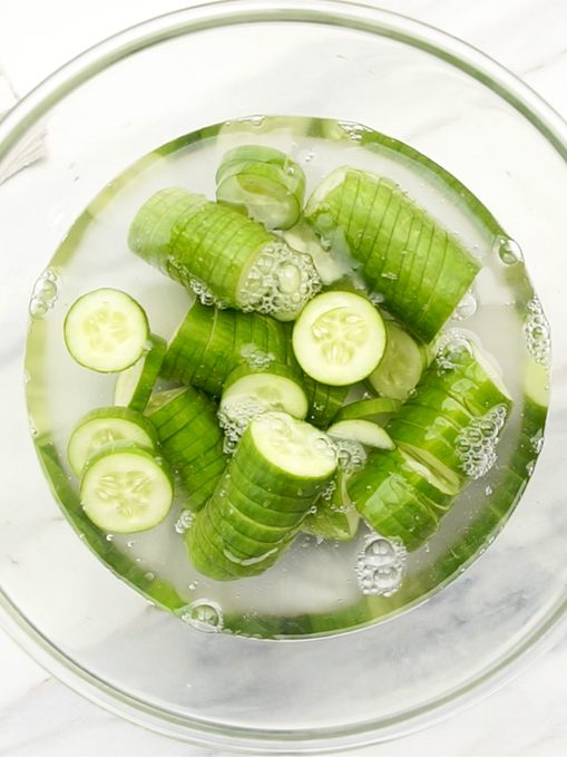 Glass bowl filled with water and salted cucumber slices, ready to soak, with ingredients for Korean cucumber salad on the side.