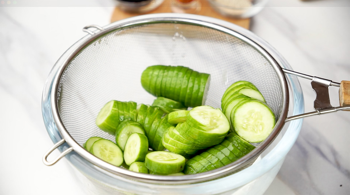 Partially sliced cucumbers draining in a fine mesh strainer over a glass bowl after soaking.
