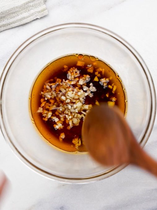 Wooden spoon stirring a mixture of soy sauce, garlic, and chili oil in a small glass bowl, with sesame seeds and garlic on the side.