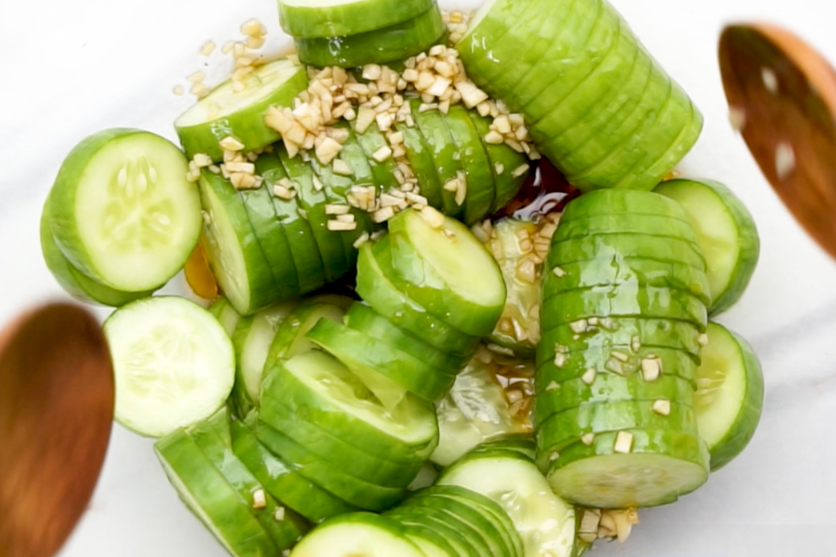 Close-up of chopped garlic soy dressing being poured over sliced cucumbers in a mixing bowl.
