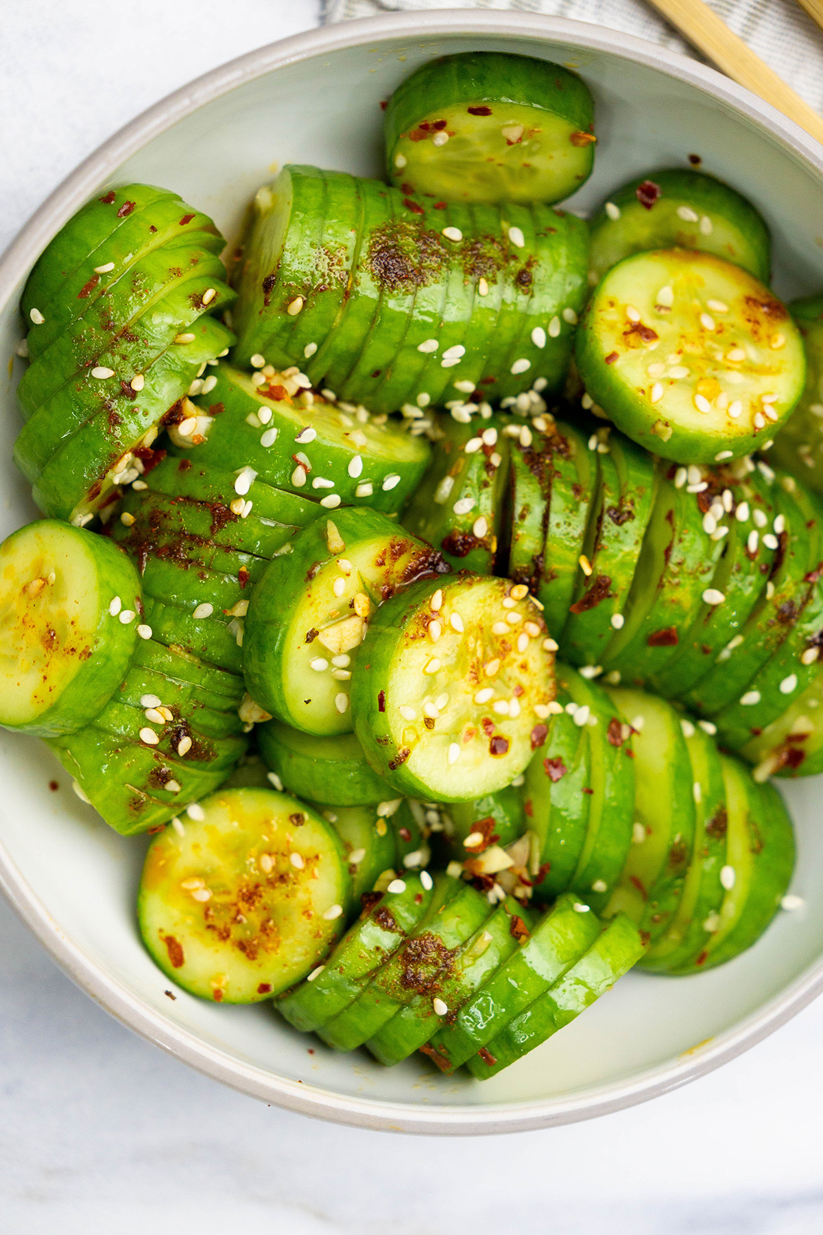Bright green cucumber slices coated in a chili-garlic dressing, sprinkled with sesame seeds, in a light gray bowl.