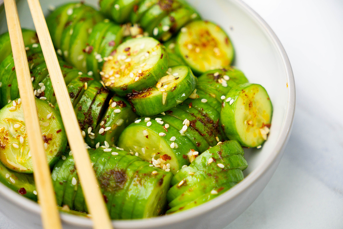 Sliced cucumbers layered in a bowl, topped with sesame seeds and chili flakes, with wooden chopsticks placed diagonally across the top.