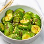Sliced cucumbers tossed in a spicy sesame dressing, garnished with sesame seeds and red pepper flakes, with chopsticks resting on side.