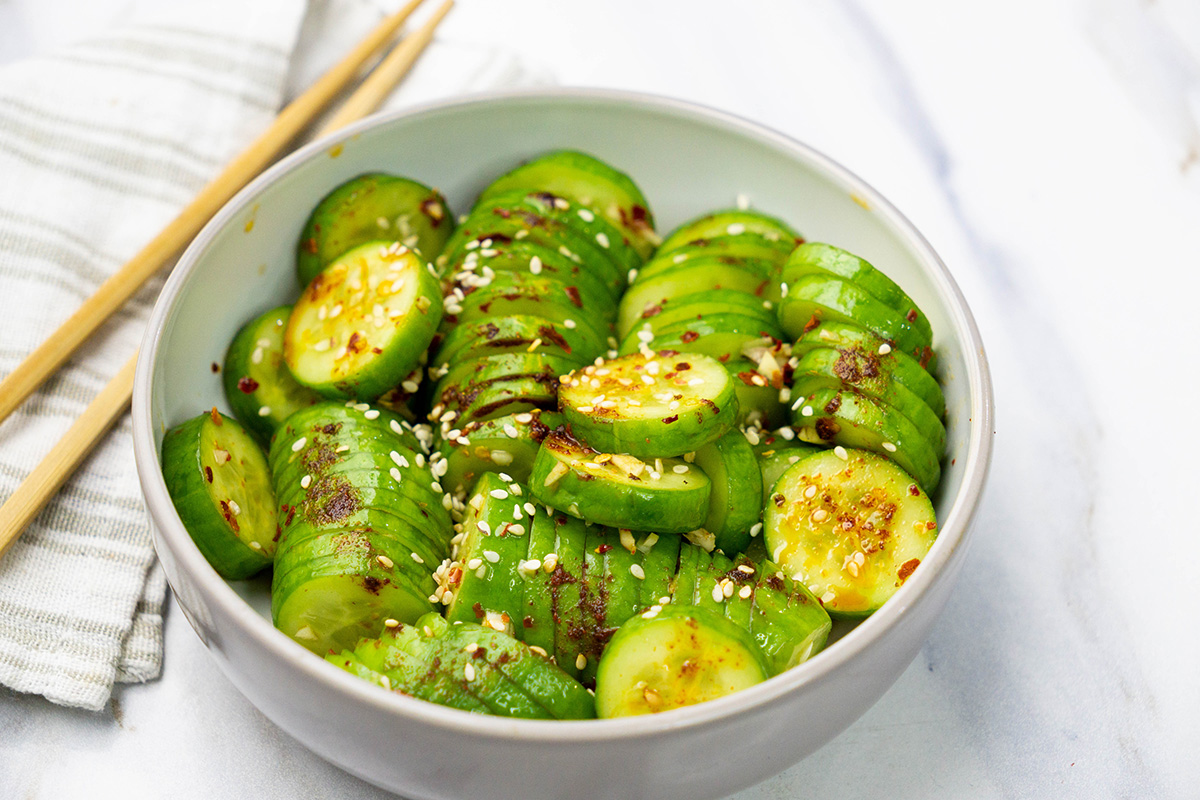 Sliced cucumbers tossed in a spicy sesame dressing, garnished with sesame seeds and red pepper flakes, with chopsticks resting on side.