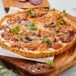 A rustic onion tart with melted cheese and browned onion slices, displayed on a wooden cutting board with scattered herbs and seasonings.