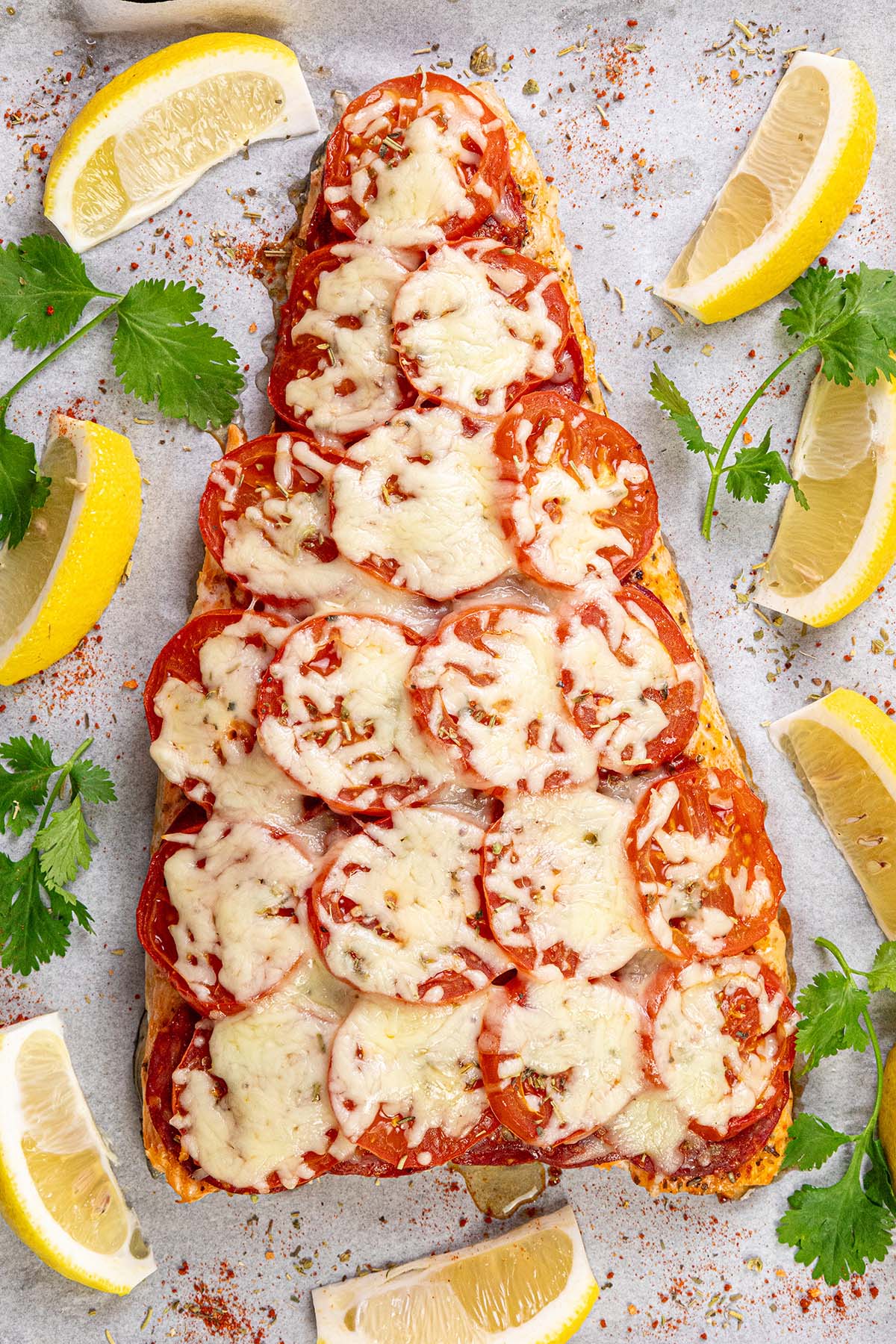 Chilean salmon on a baking sheet topped with chorizo, tomato and melted mozzarella cheese garnished with lemon wedges and parsley.