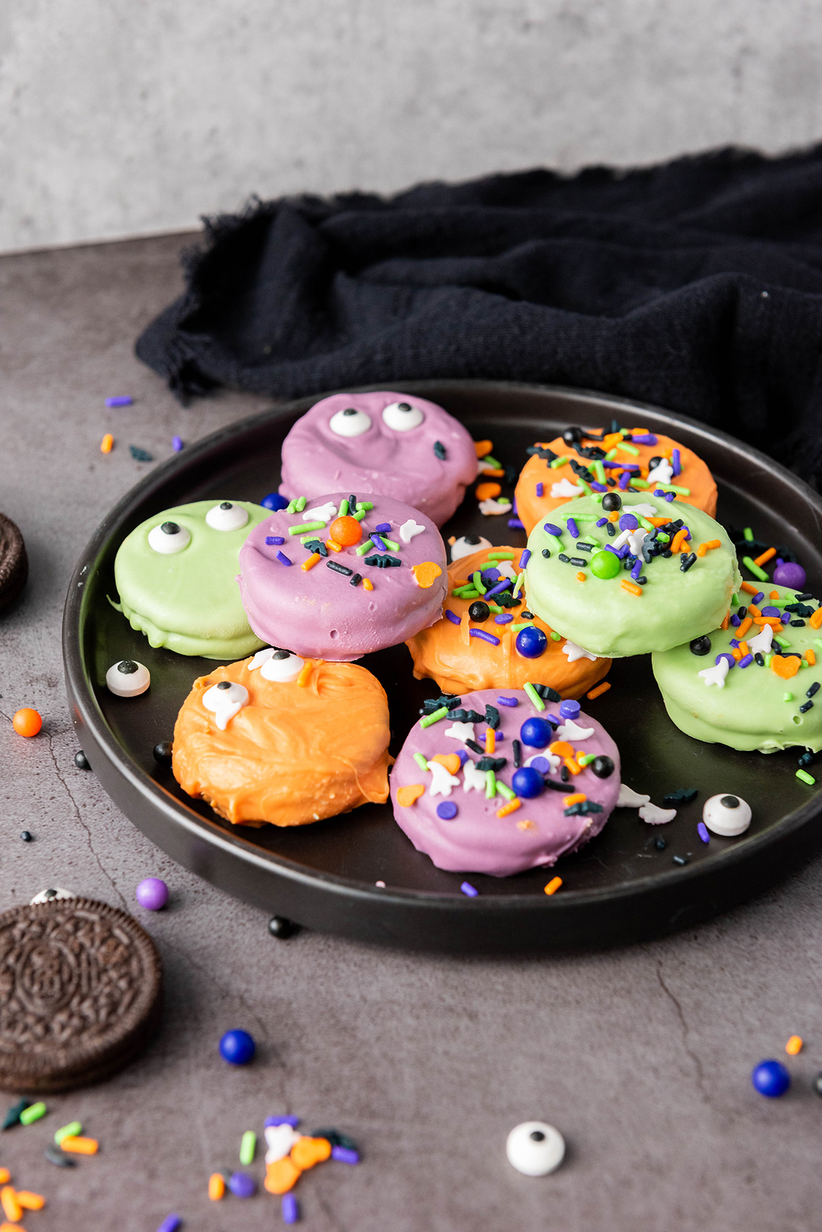These chocolate covered Halloween Oreos are the easiest spooky treat! Dip, decorate, and serve a colorful no-bake treat that’s just as fun to decorate as it is to eat! Black plate filled with colorful Halloween themed chocolate covered Oreos, sprinkles scattered around.