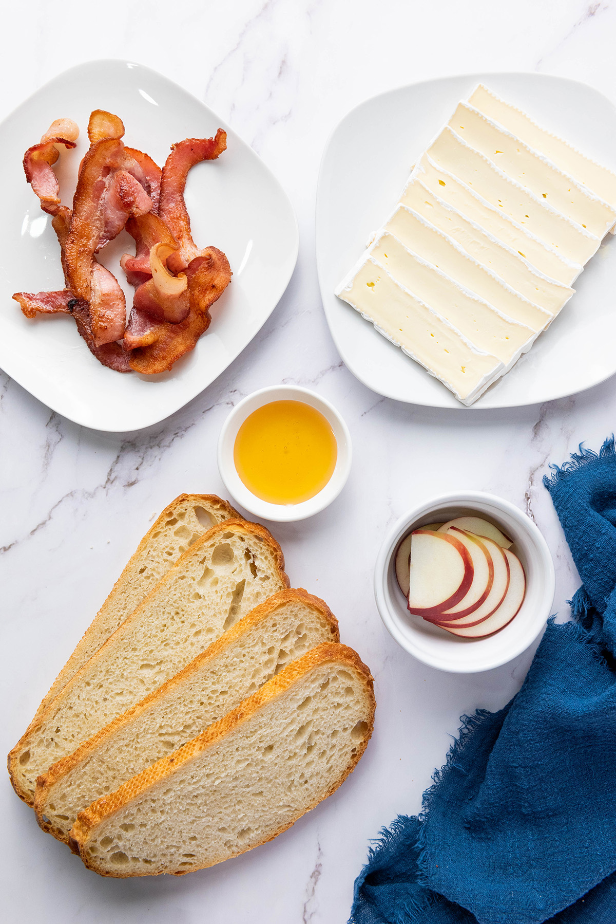 Ingredients for apple bacon and brie panini with sourdough bread, crispy bacon, brie cheese slices, apple slices, and honey on a marble surface.