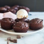 Four brownie cakes on a plate with one topped with a scoop of vanilla ice cream, pink cloth napkin and cooling rack in the background.