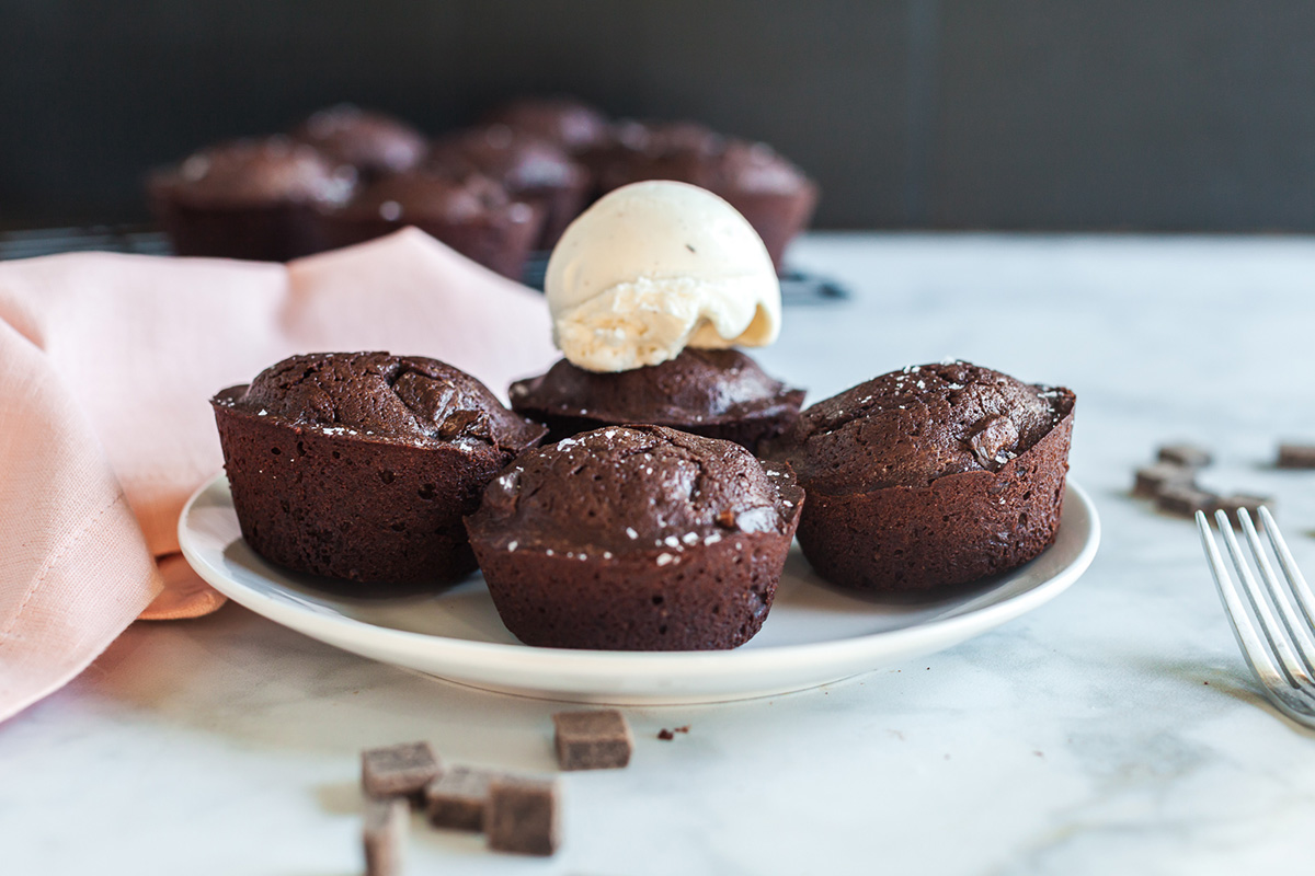 Part brownie, part cake, these brownie cakes are rich like a fudgy brownie but baked in cupcake form and made completely from scratch. Four brownie cakes on a plate with one topped with a scoop of vanilla ice cream, pink cloth napkin and cooling rack in the background.