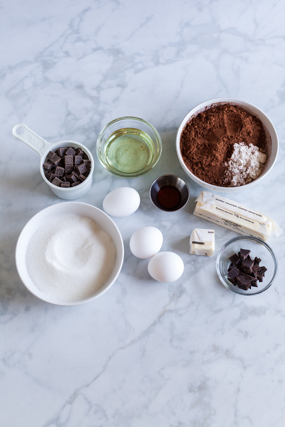 Part brownie, part cake, these brownie cakes are rich like a fudgy brownie but baked in cupcake form and made completely from scratch. Overhead view of brownie cake ingredients on a marble surface, including eggs, sugar, cocoa powder, flour, butter, oil, vanilla, and chocolate chunks.