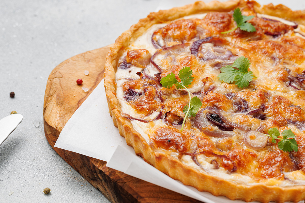 Flaky pastry, sweet caramelized onions and a creamy egg and Gruyere cheese filling make this onion tart both flavorful and irresistible to serve as an appetizer, dinner or even brunch. Caramelized onion tart with a golden, flaky crust, sitting on parchment paper over a wooden board and sprinkled with herbs.