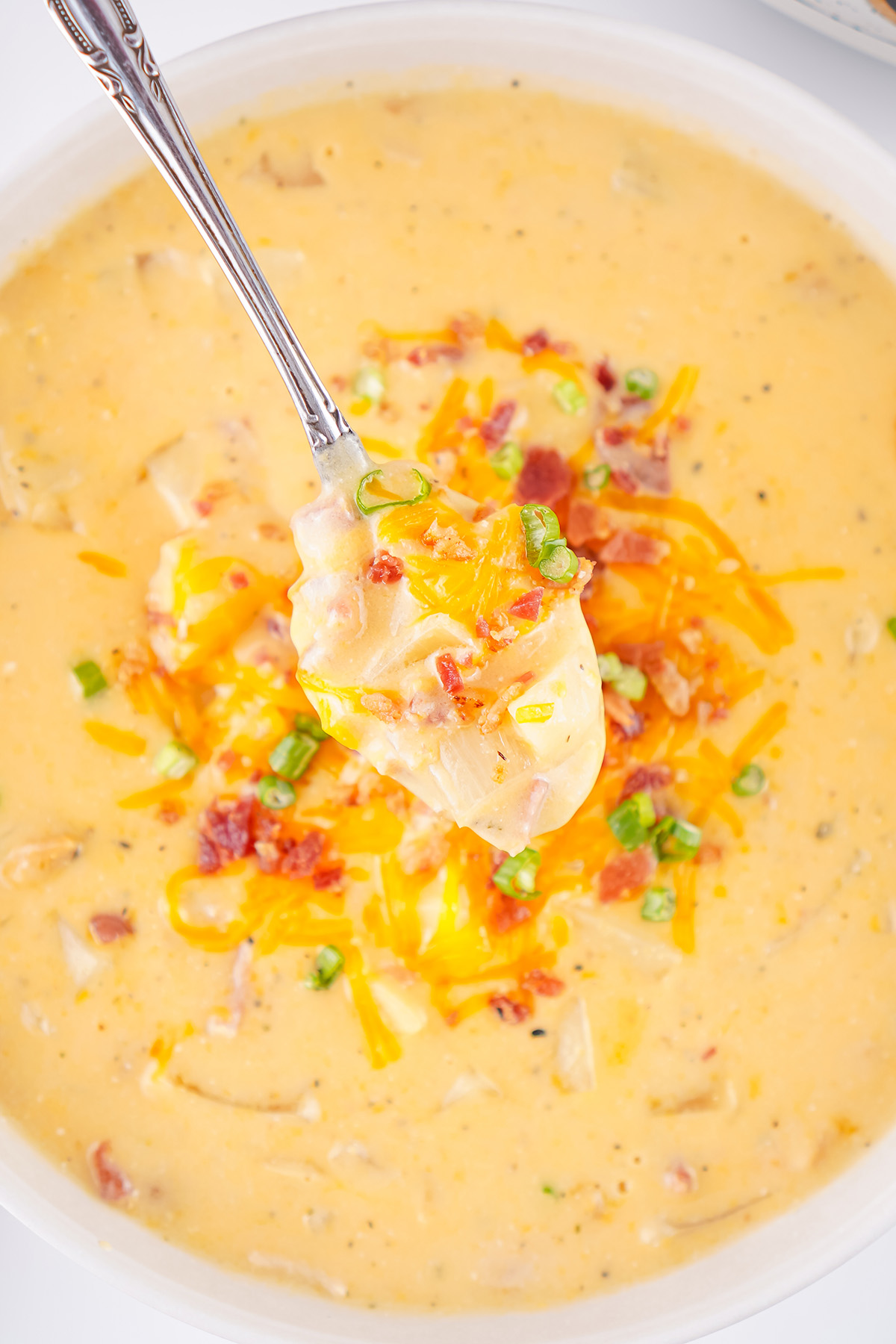 Silver spoon scooping cheesy crock pot crack potato soup topped with crispy bacon and sliced green onions in a white bowl.